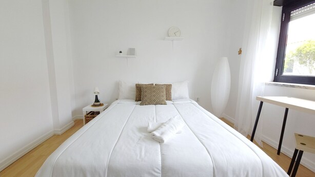 A neatly made bed with white bedding and a folded towel, a bedside table with a lamp, a small plant, and a wall clock. - Freedom Surf House (Peniche)