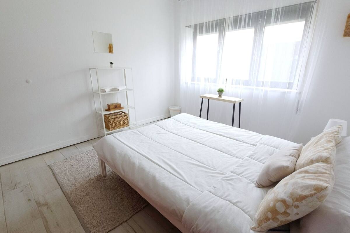A minimalist bedroom with a large bed, a small table, a shelf unit, and a window with sheer curtains.