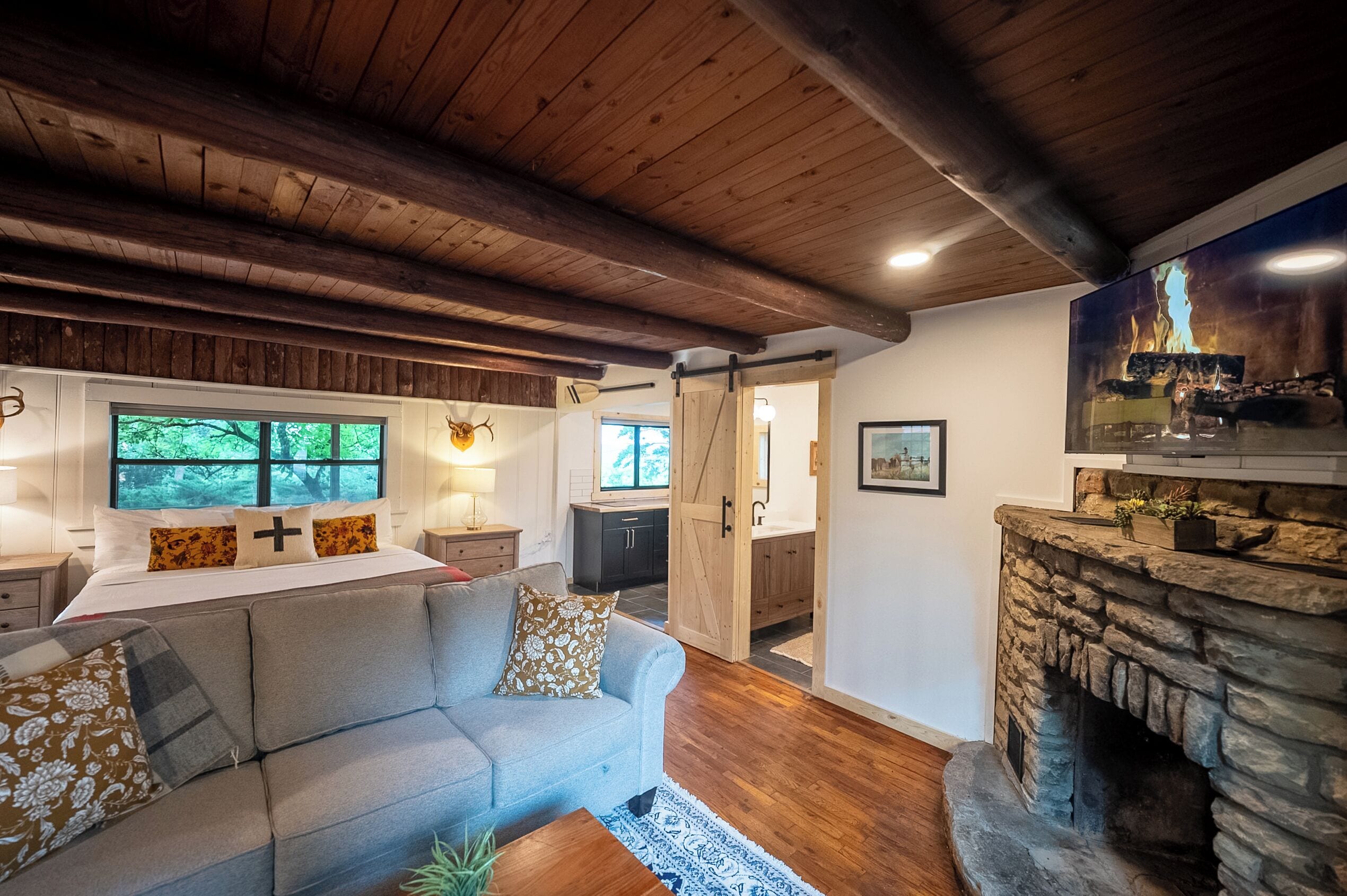 A cozy living room with a stone fireplace, a sofa with patterned cushions, a bed with a view of greenery, and a kitchen area visible through an open door.