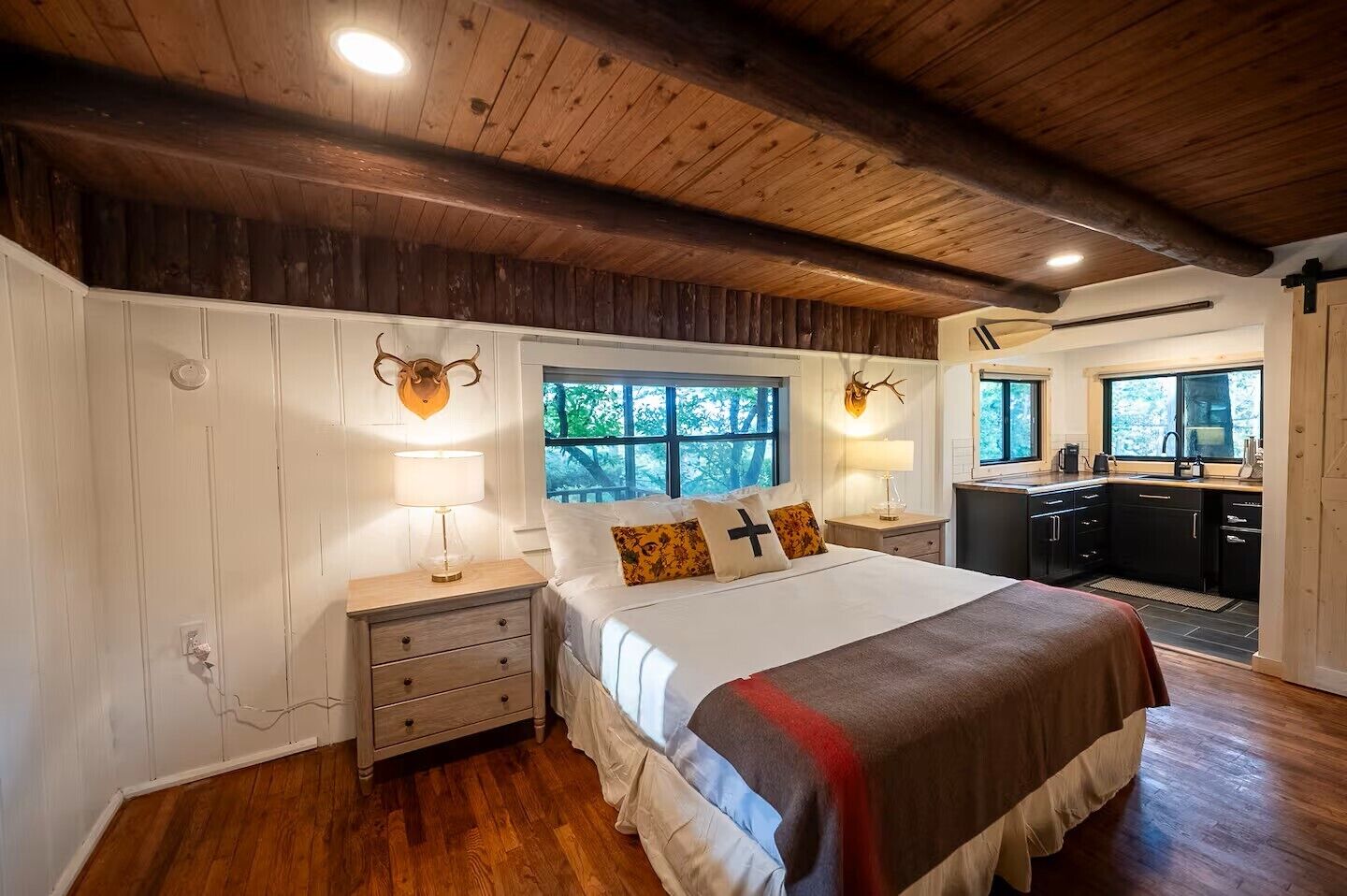 Un dormitorio con techo de madera, una cama con manta roja, una mesita de noche con lámpara y vistas al exterior a través de una ventana.