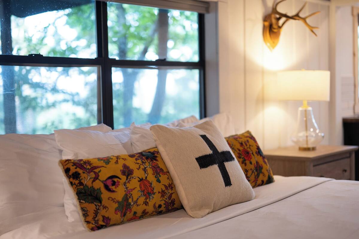 A bed with white and yellow floral pillows, a black cross pillow, and a deer head wall decoration.