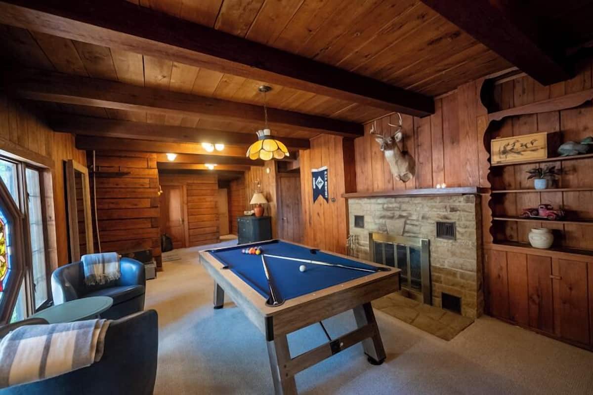 A wooden interior with a pool table, a fireplace, and a deer head mounted on the wall.