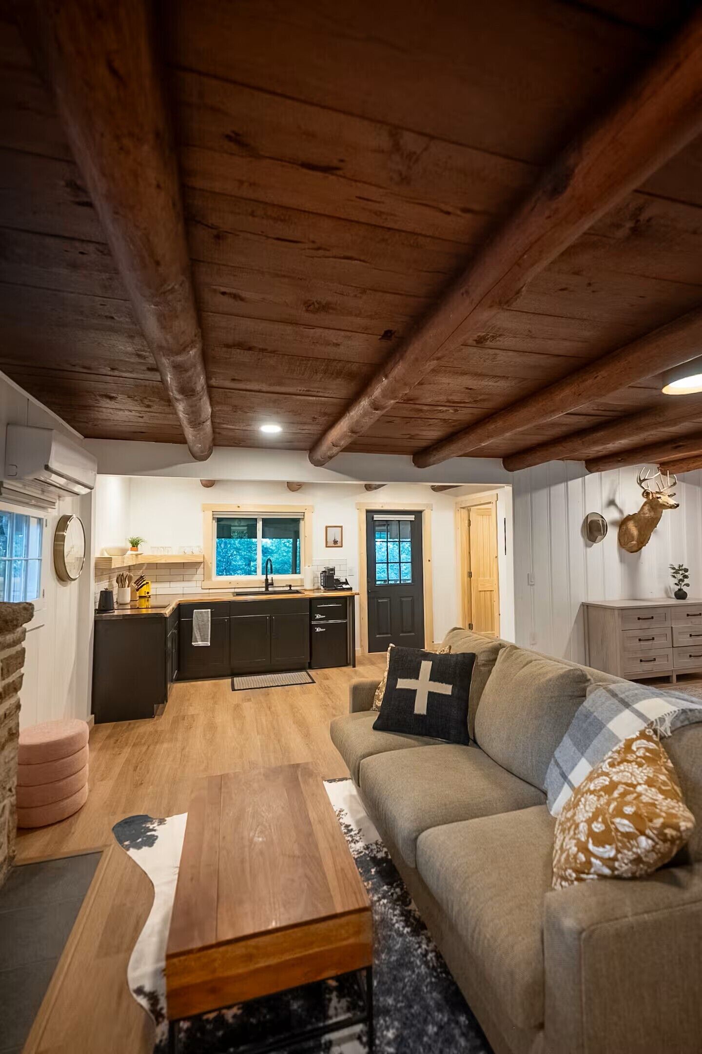 Ein gemütliches Interieur mit Holzbalkendecke, einer Küche mit Fenster, einem Sofa mit Kreuzkissen und einem Hirschgeweih an der Wand.