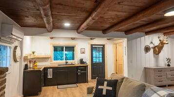 Ein gemütliches Interieur mit Holzbalkendecke, einer Küche mit Fenster, einem Sofa mit Kreuzkissen und einem Hirschgeweih an der Wand.