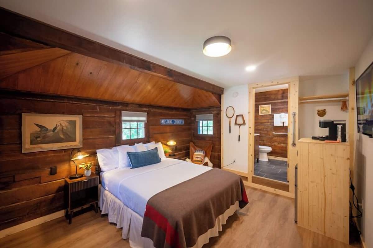 A wooden cabin-style bedroom with a bed, bedside lamps, a framed picture, and a wooden cabinet.