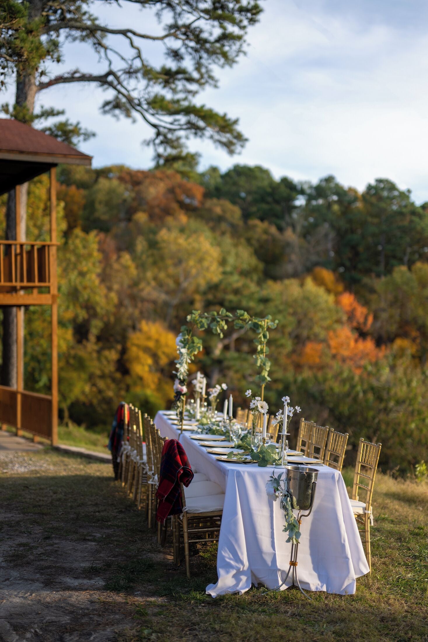 An outdoor dining setup with white tablecloths and gold chairs, set against a backdrop of autumn foliage and a wooden structure.