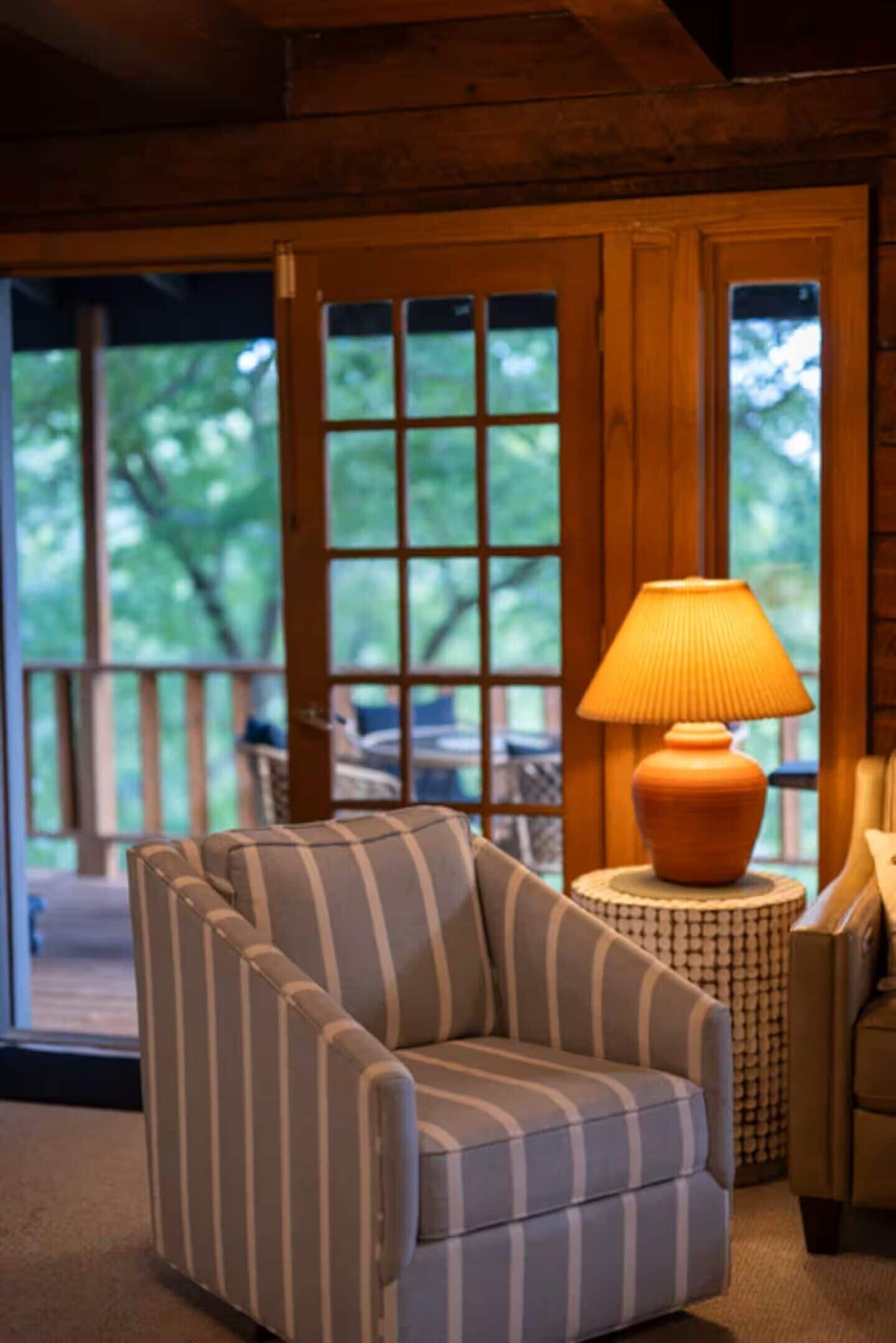 A room with a striped armchair, a round table with a lamp, and a view through large windows.