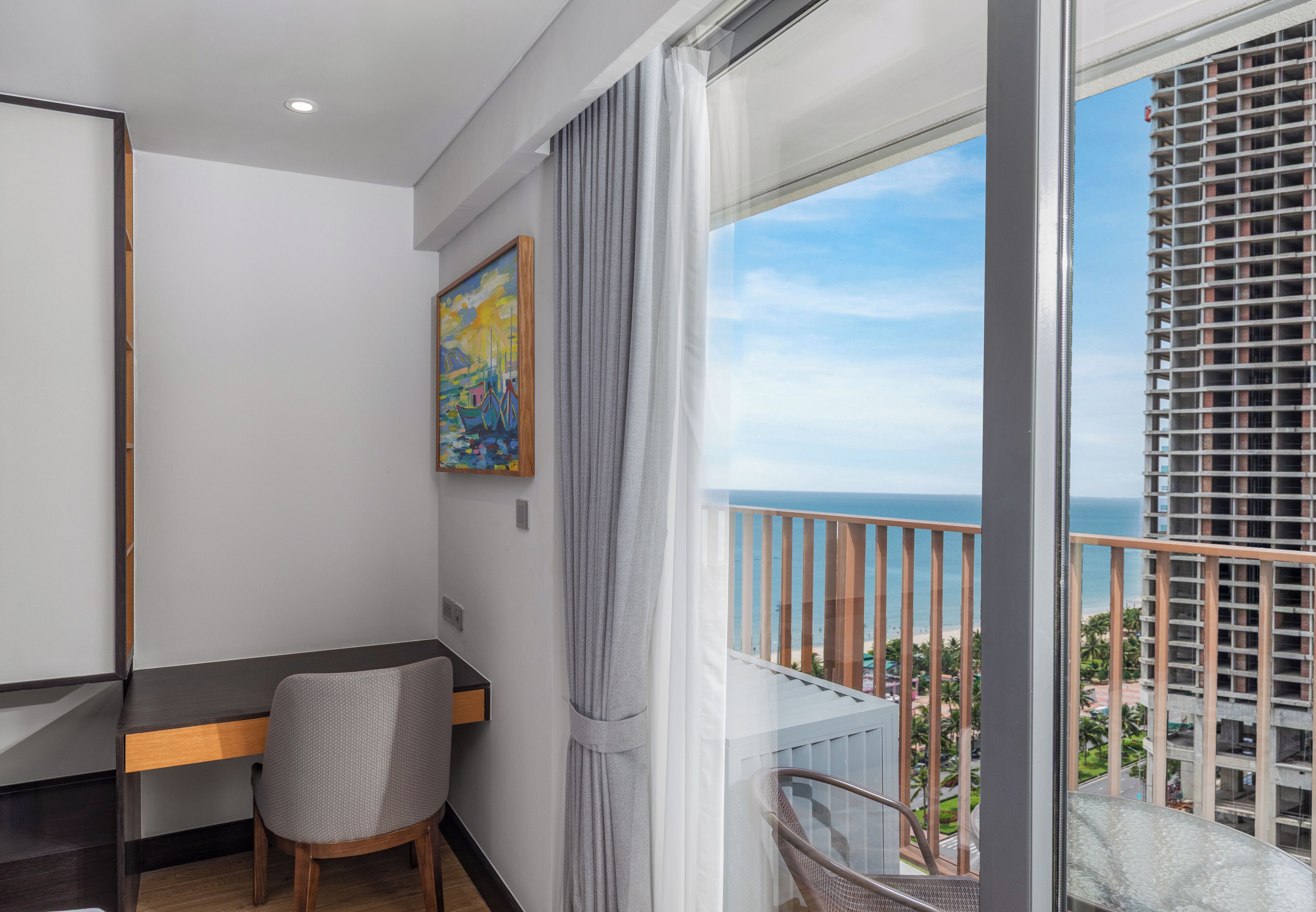 A modern hotel room with a balcony, a desk, a chair, and a painting on the wall.
