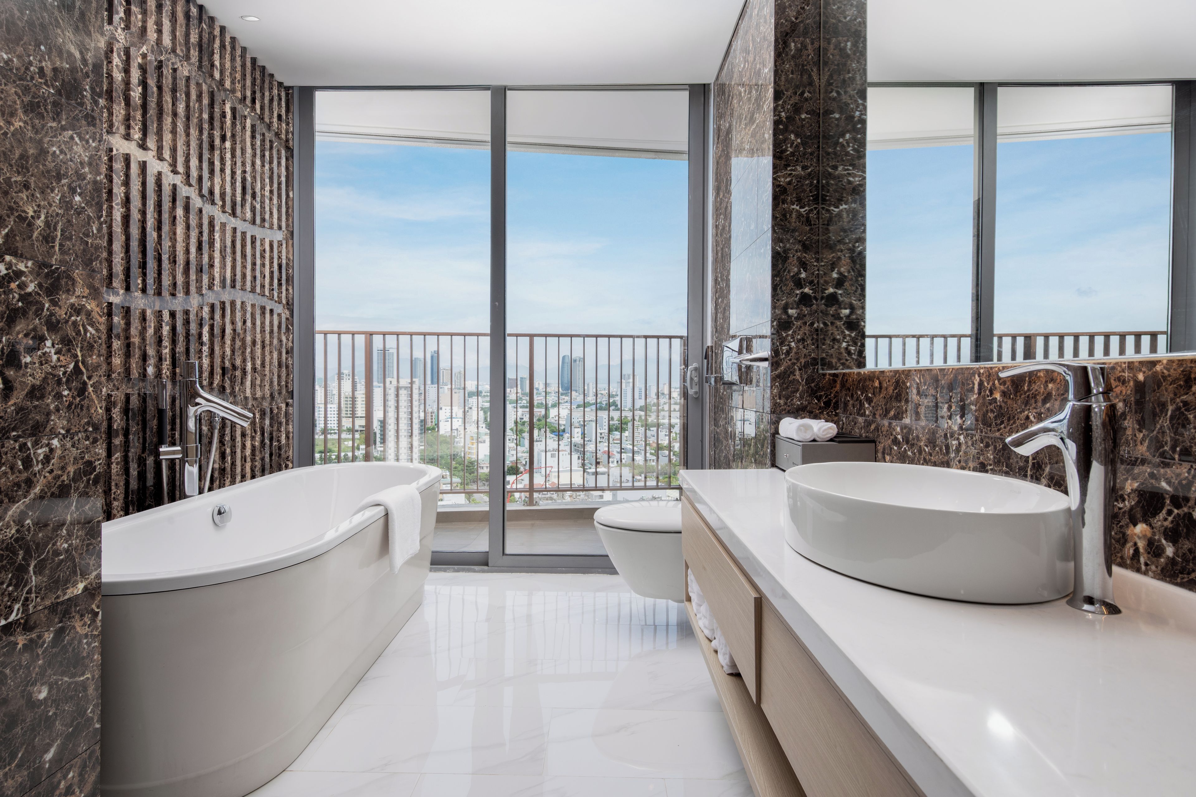 Une salle de bain moderne avec une grande baignoire, un lavabo rond et des toilettes. Elle est dotée d’une grande fenêtre offrant une vue sur la ville.