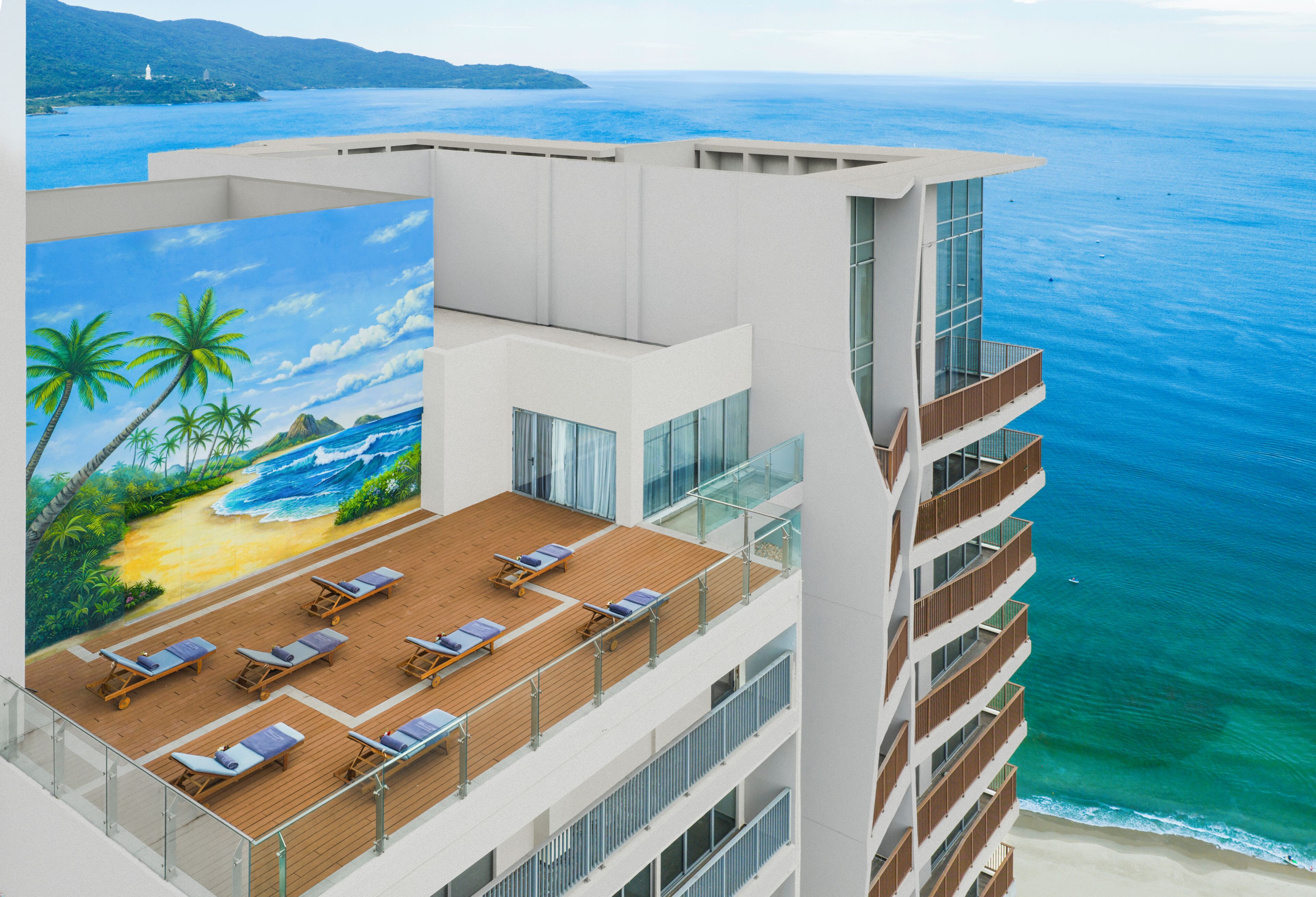 A multi-story hotel with a rooftop deck featuring sun loungers and a mural of a beach scene.
