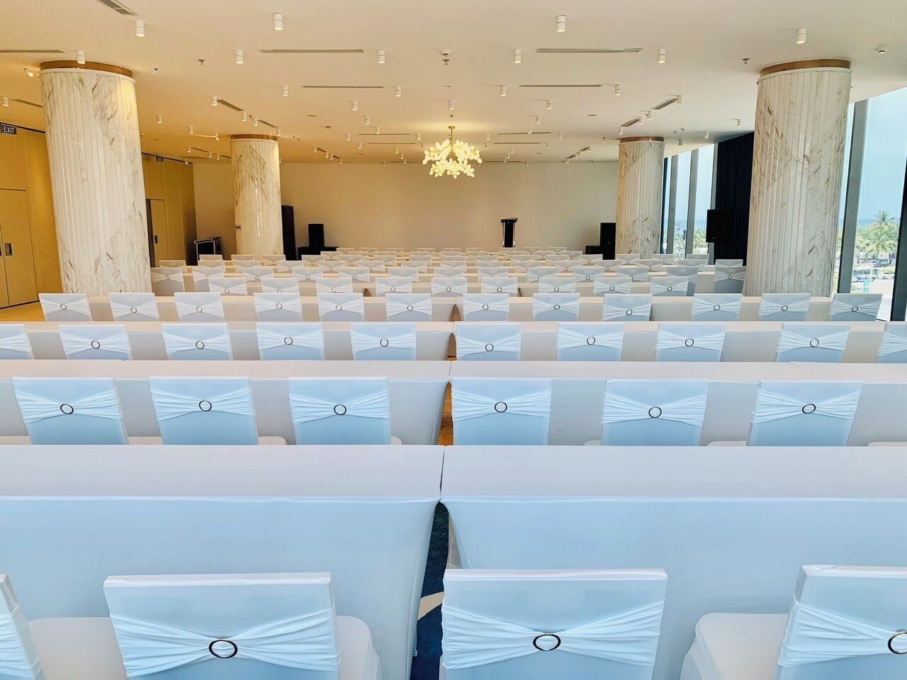 Un salón de conferencias con filas de mesas y sillas blancas, pilares de mármol y una araña.