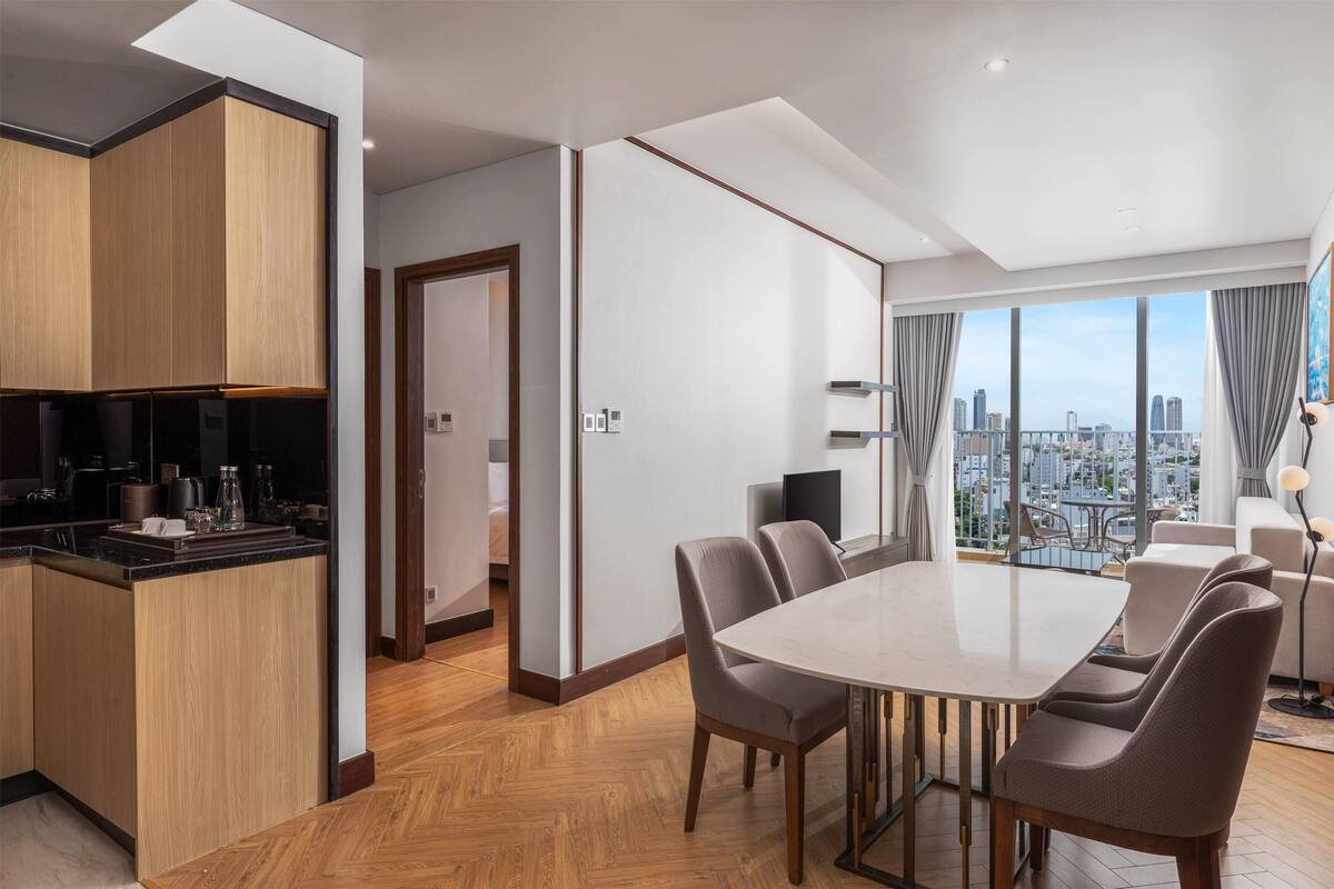 Una habitación de hotel moderna con una amplia mesa de comedor, asientos confortables, una zona de cocina y vistas a la ciudad a través de grandes ventanales.
