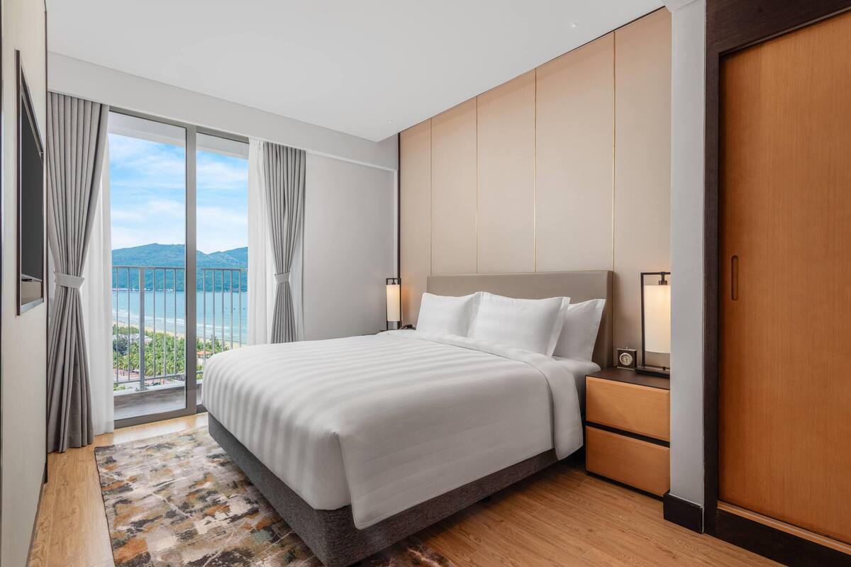 Habitación de hotel con una cama grande, una mesita de noche, un armario de madera y vistas al mar.