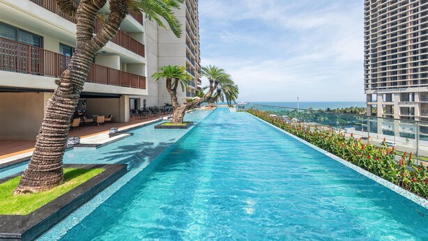 A long swimming pool with a clear view of the ocean, surrounded by modern buildings and palm trees. - Wyndham Soleil Danang - Free Schedule Airport Shuttle (Da Nang)