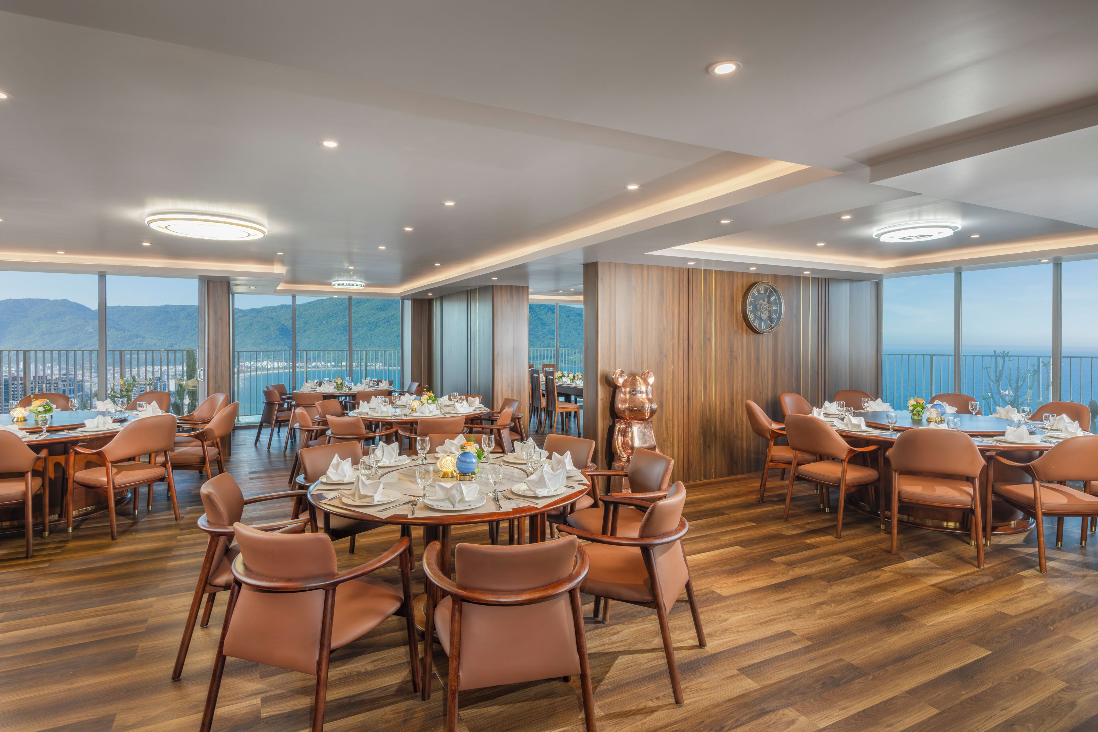A spacious dining area with multiple tables set for dining, large windows offering a view of the city and mountains, and a modern ceiling design.