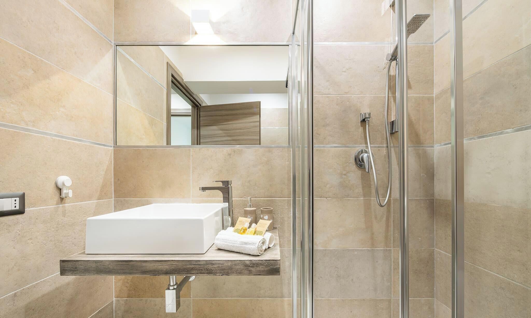 Un baño moderno con cabina de ducha acristalada, lavamanos blanco y estante de madera con artículos de aseo.