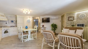 A well-lit kitchen with white cabinets, a dining table with chairs, a TV mounted on the wall, and a bed with white bedding. - Villa Rachele ground floor apt, private pool (Porto Pinetto)