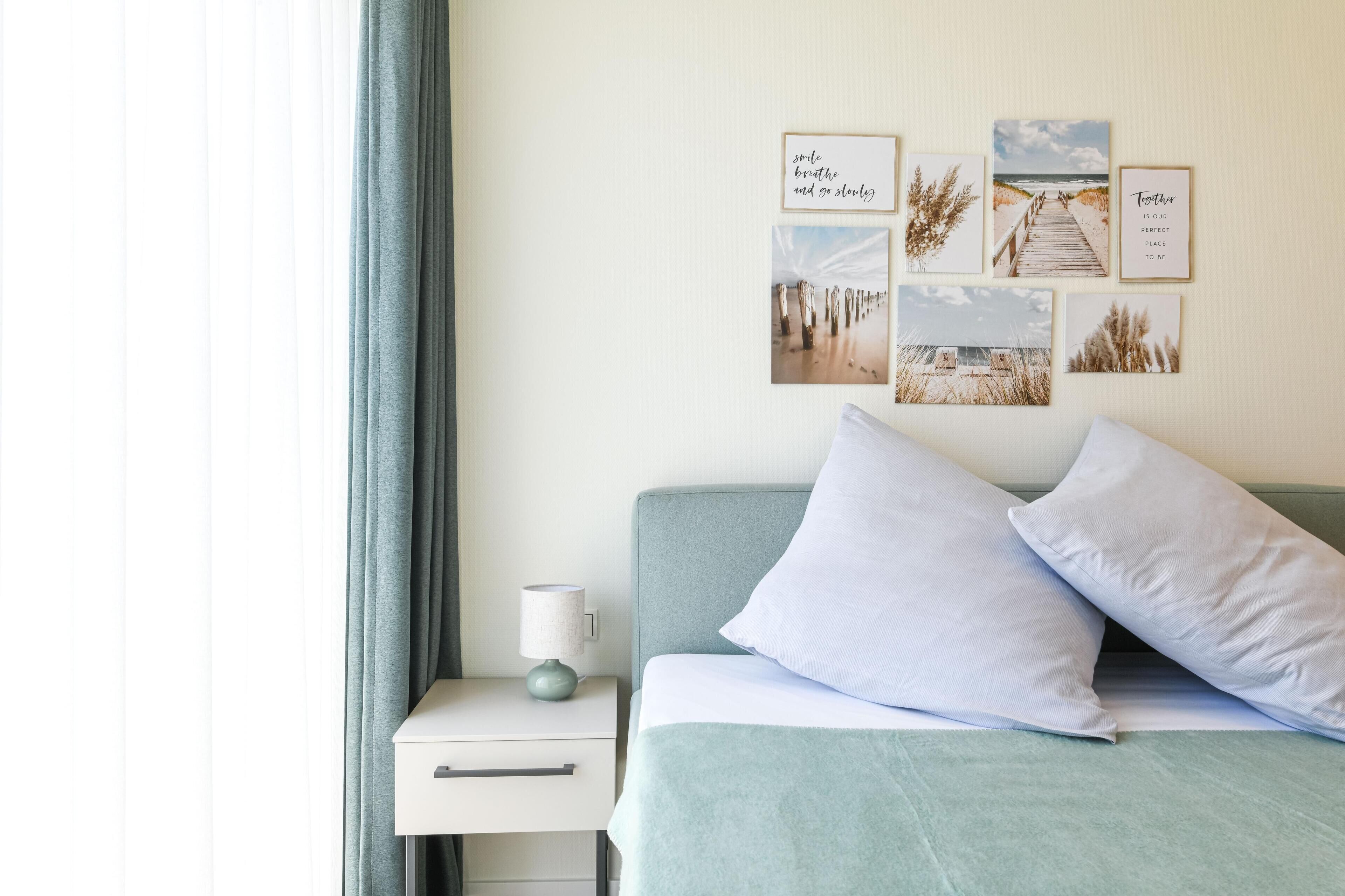 Un dormitorio ordenado con una cama, almohadas, una mesita de noche con lámpara y una pared decorada con cuadros.