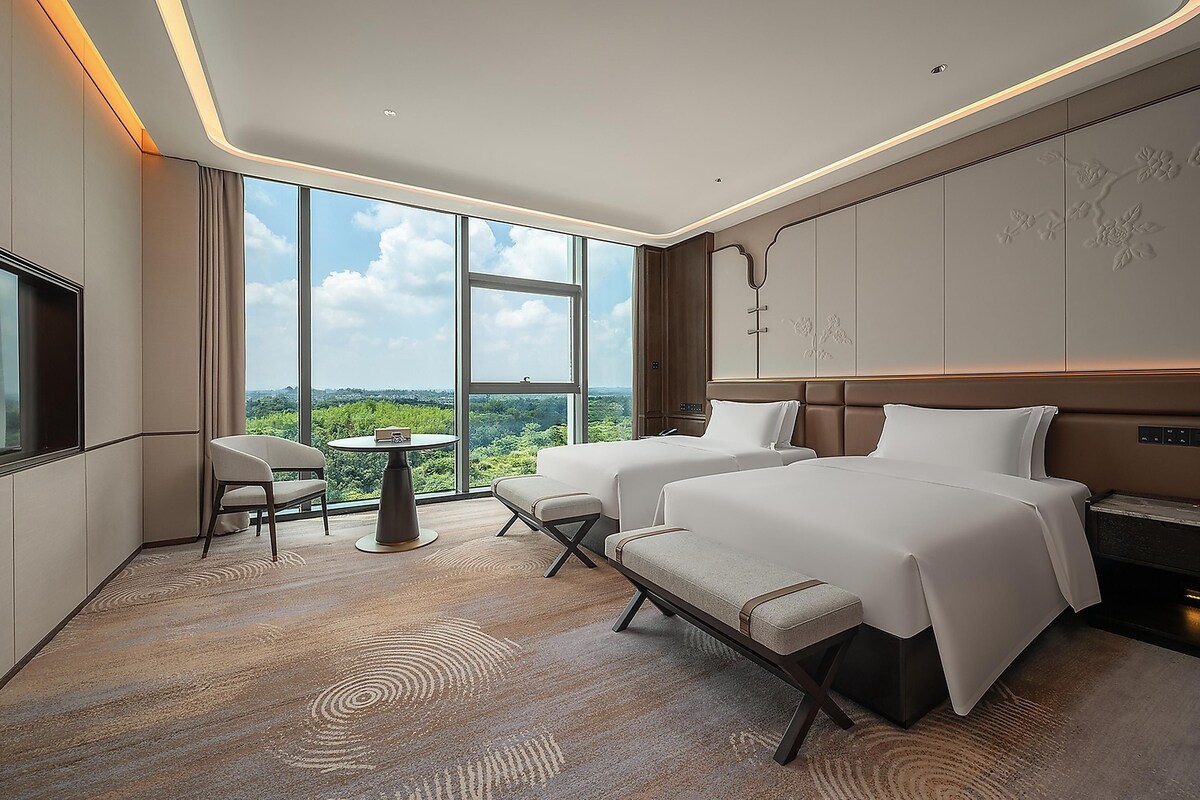 Habitación de hotel con dos camas, zona de estar y un amplio ventanal con vistas a la ciudad.