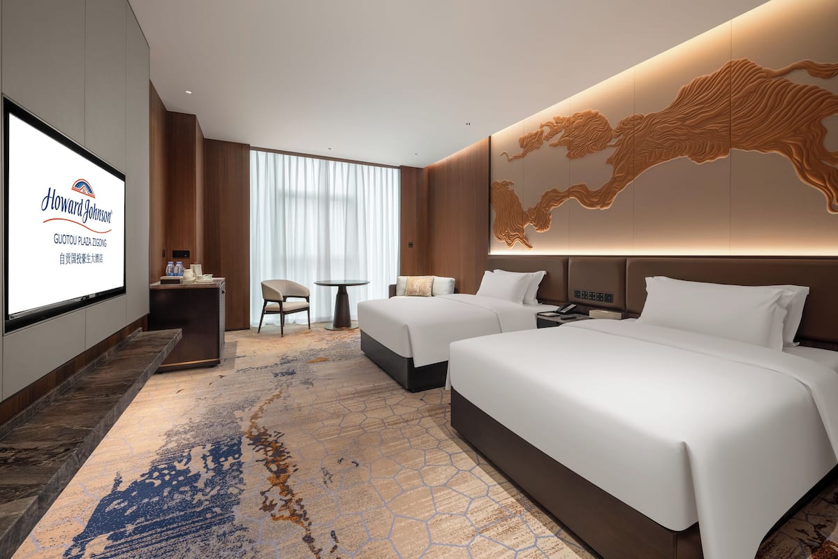 A hotel room with two beds, a desk, a TV screen displaying the hotel name, and a large wall mural.