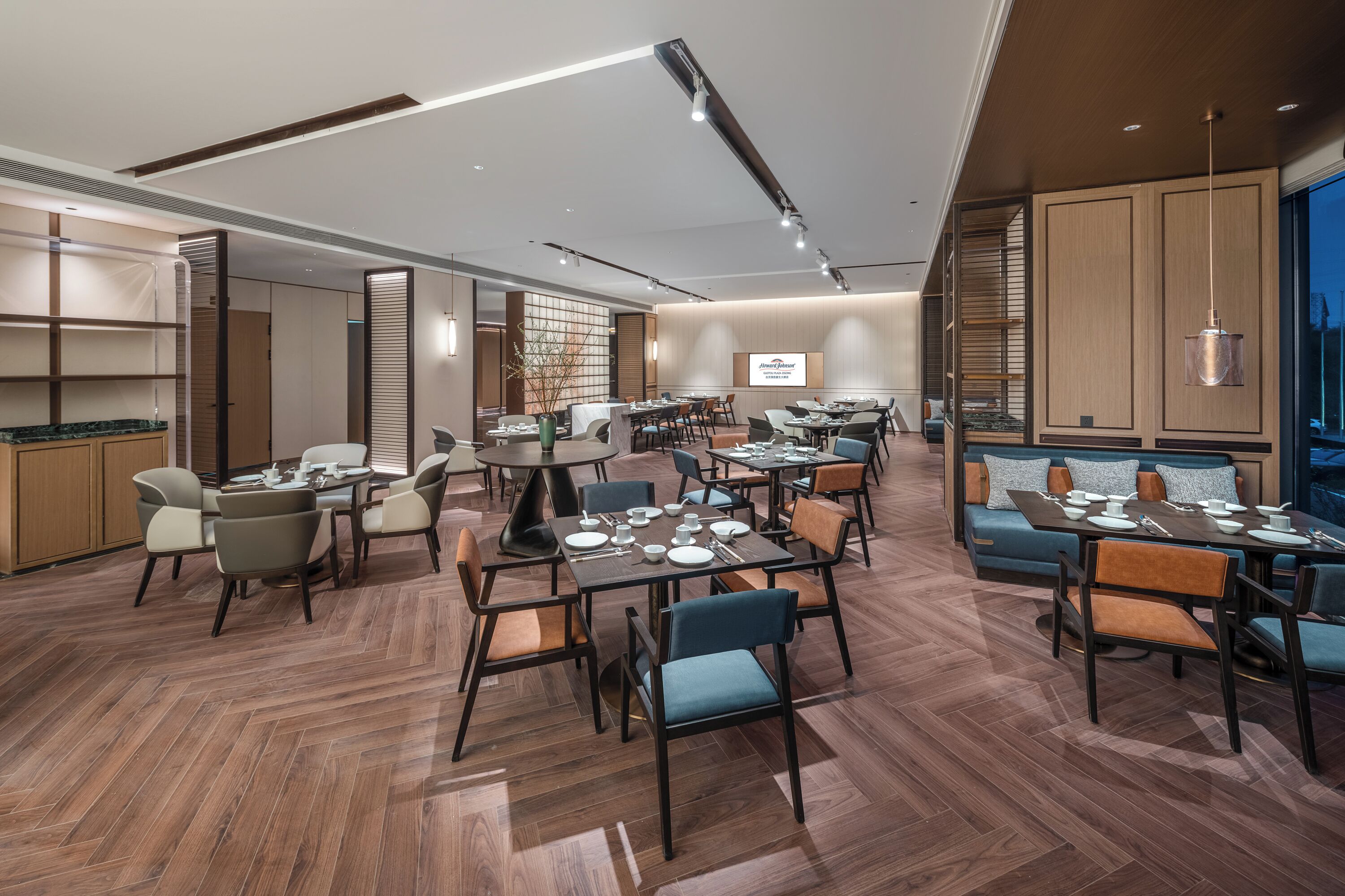 A modern dining area with multiple tables and chairs, wooden flooring, and a large screen displaying the hotel name.
