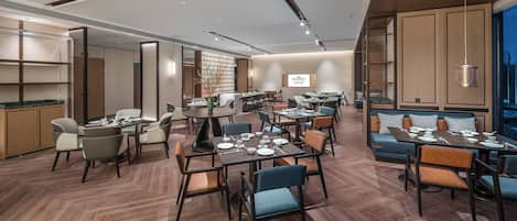 A modern dining area with multiple tables and chairs, wooden flooring, and a large screen displaying the hotel name.