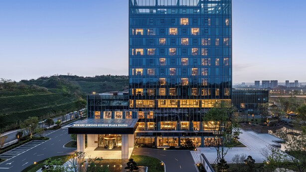 A modern hotel with a glass facade, a well-lit entrance, and a landscaped driveway.