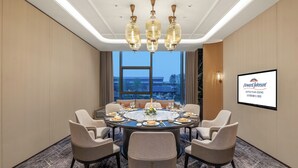 A conference room with a round table, six chairs, and a chandelier. A screen displays the hotel name and location.