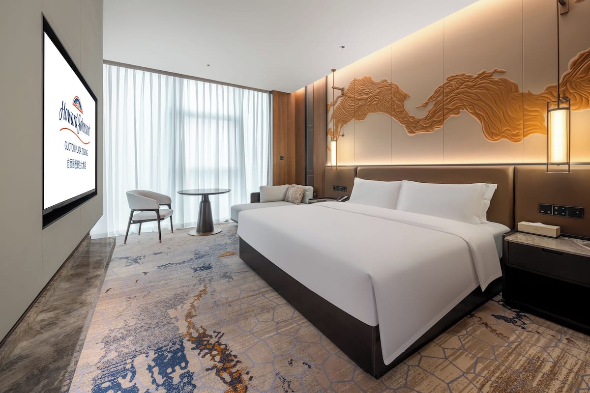 A modern hotel room with a large bed, a desk, and a television displaying the hotel name.