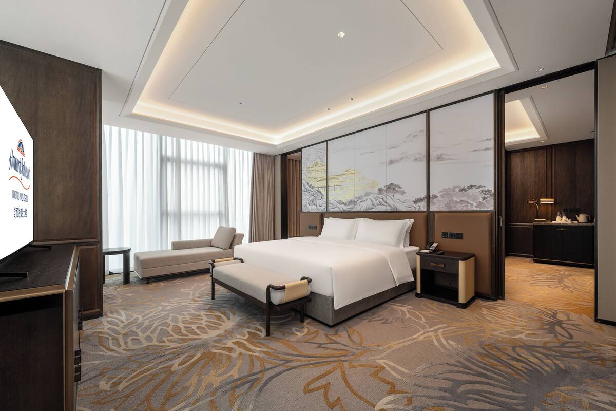 A modern hotel room with a large bed, a desk, a sofa, and a view of a mountain landscape through the window.