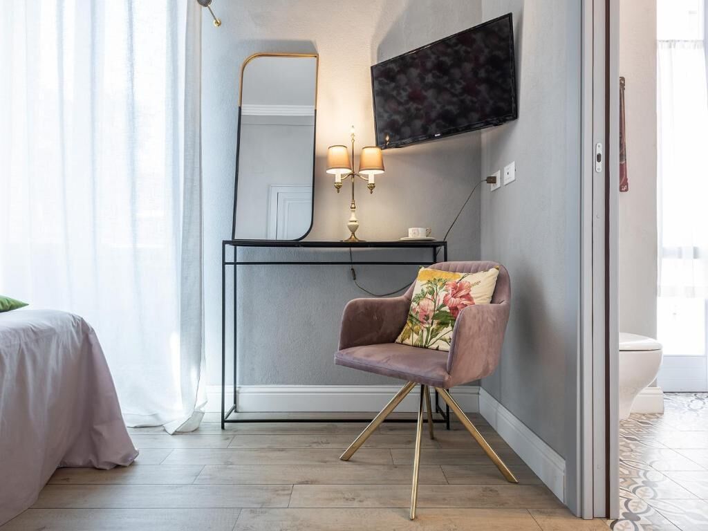 A modern bedroom with a bed, a vanity with a mirror, a chair with a floral pillow, and a TV mounted on the wall.