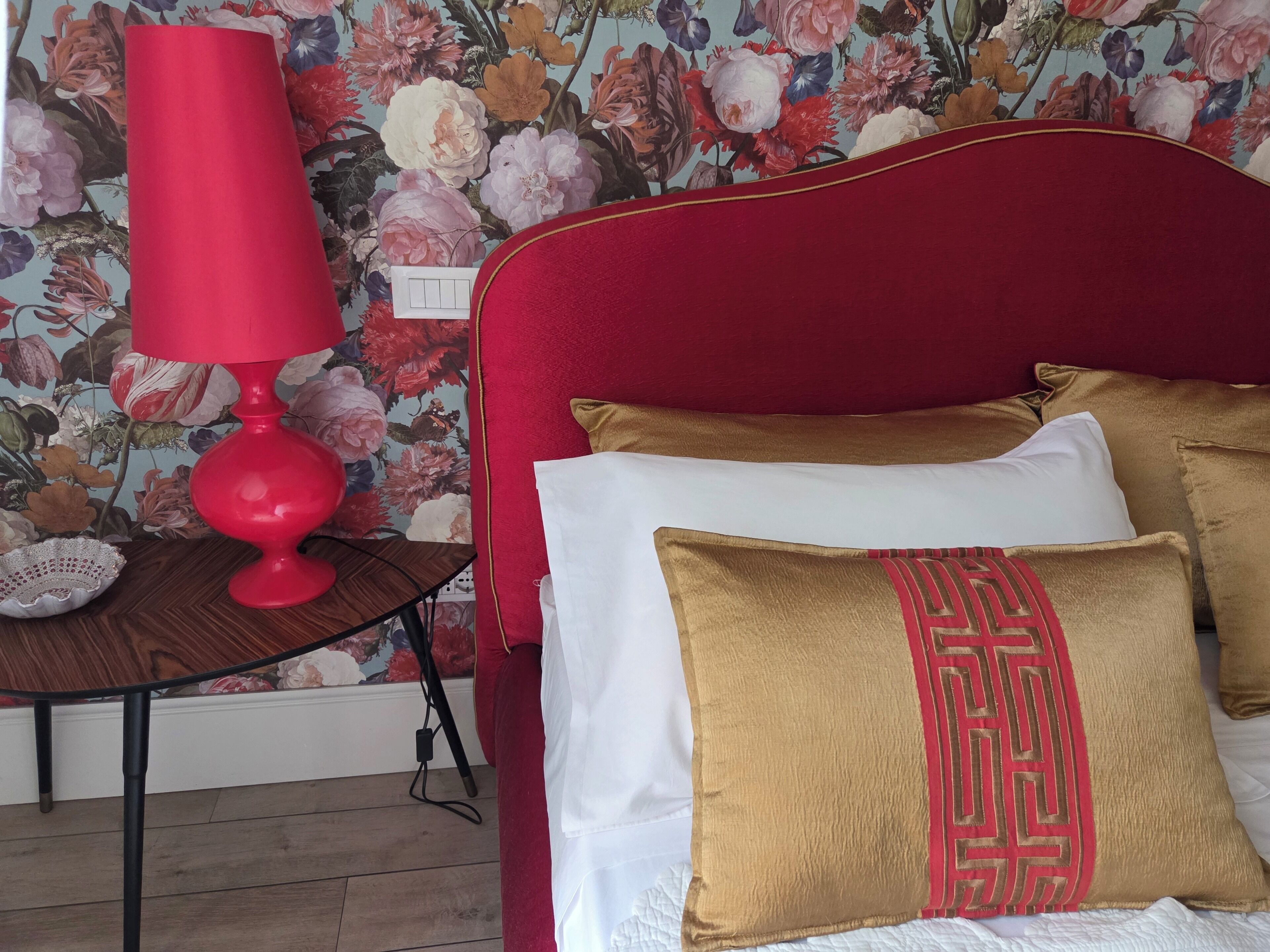 A bed with red headboard, white and gold pillows, and a red lamp on a wooden nightstand.