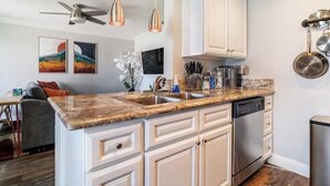 A kitchen with white cabinets, a granite countertop, a dishwasher, and a ceiling fan. - Charming & Cozy Condo • Pool-Spa & Pac-Man Arcade (Indio)