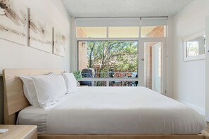 A bedroom with a bed, a nightstand, a window with a view of the outside, and a door leading to another room.
