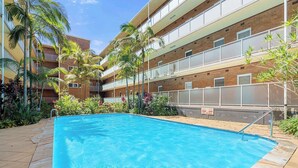 A swimming pool with a tiled deck, surrounded by palm trees and a multi-story building with balconies. - Spacious and Idyllic Unit in Little Manly (Manly)