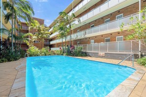 A swimming pool with a tiled deck, surrounded by palm trees and a multi-story building with balconies.