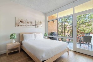 A bedroom with a large window, a bed, a nightstand, and artwork on the wall.