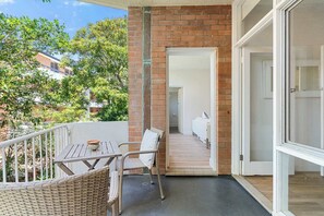 A balcony with wicker furniture, a door leading to an interior space, and a view of trees and buildings.