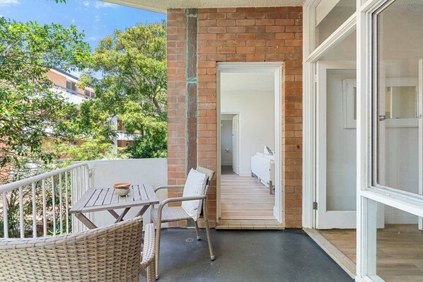 A balcony with wicker furniture, a door leading to an interior space, and a view of trees and buildings. - Spacious and Idyllic Unit in Little Manly (Manly)
