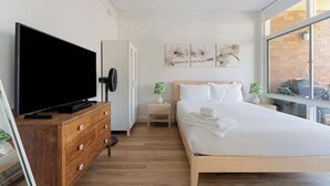 A modern hotel room with a large flat-screen TV, wooden dresser, a bed with white linens, a small side table, and artwork on the wall. - Spacious and Idyllic Unit in Little Manly (Manly)