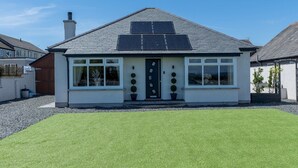 A single-story house with a dark roof, white walls, and a front door with a decorative glass panel. There are two solar panels on the roof, and the property is surrounded by a well-maintained lawn.