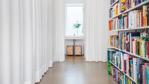 Ein heller, gut beleuchteter Raum mit einem großen Bücherregal voller Bücher, einem Fenster mit weißen Vorhängen und einem Holzschrank.