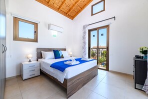 Ein Schlafzimmer mit Holzbalkendecke, einem Bett mit weißer Bettwäsche und blauen Akzenten, einem Nachttisch, einer Kommode und einer Schiebetür zu einem Balkon.