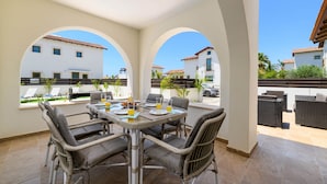 A patio with a table set for four, surrounded by wicker chairs, overlooking a pool area with lounge chairs and a building with a tiled roof. - Kymma Villa 12 in Ayia Napa (Sotira)