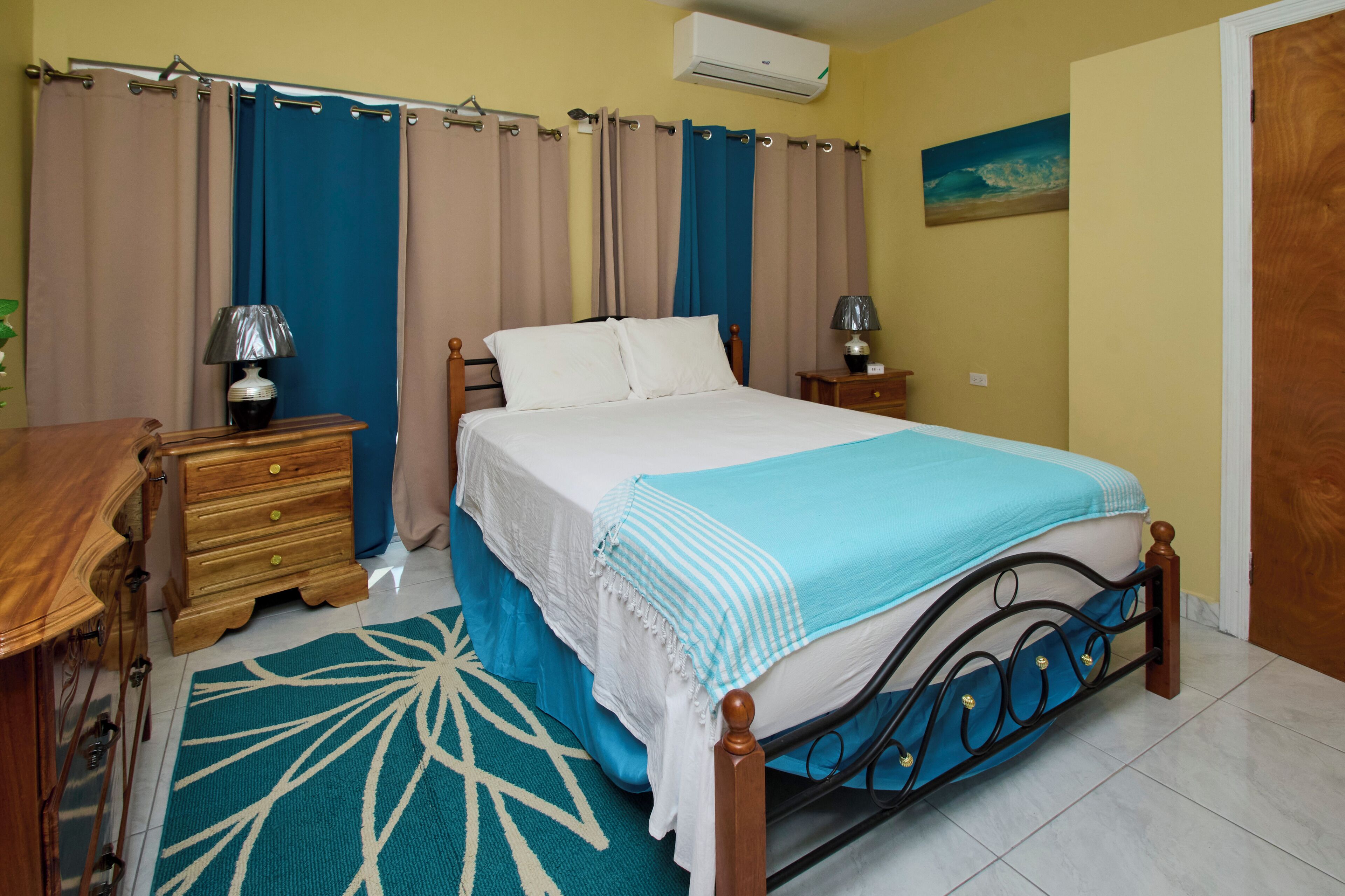 Une chambre à coucher avec un lit, des tables de chevet, une commode en bois, un tapis à motifs bleus et blancs et un tableau accroché au mur.
