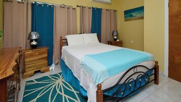 Une chambre à coucher avec un lit, des tables de chevet, une commode en bois, un tapis à motifs bleus et blancs et un tableau accroché au mur.