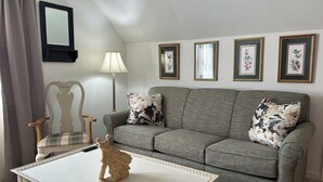 Un salon avec un canapé gris, deux coussins à motifs, une table basse blanche agrémentée d’un objet décoratif, une chaise en bois, une lampe et quatre tableaux encadrés accrochés au mur.