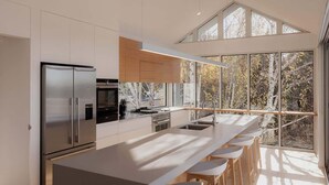 Ett modernt kök med en stor köksö, rostfria vitvaror och utsikt över träd genom stora fönster.