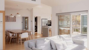 Ett modernt vardagsrum med en soffa, ett matbord och stolar. Det finns ett köksområde med ett kylskåp och en mikrovågsugn. Rummet har stora fönster och utsikt över omgivningen.