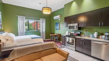 A room with a bed, a sofa, a dining table, a kitchen with a stove, and an air conditioning unit.